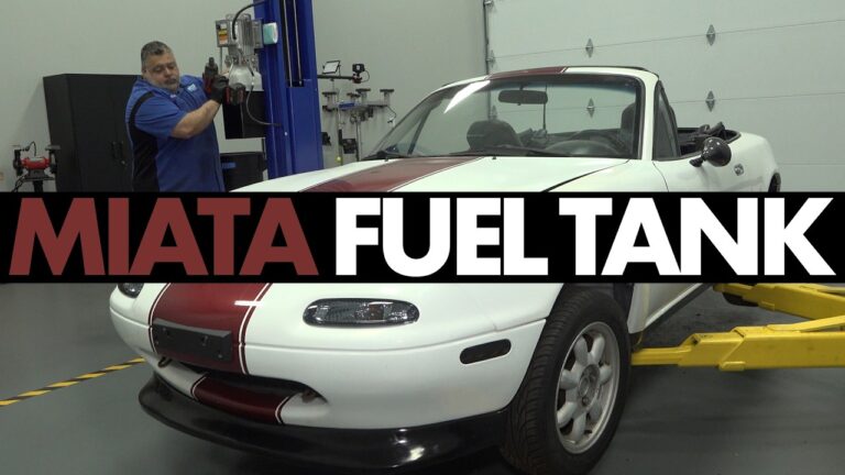 A mechanic working on the underside of a Mazda Miata to remove the rear subframe for fuel tank access.